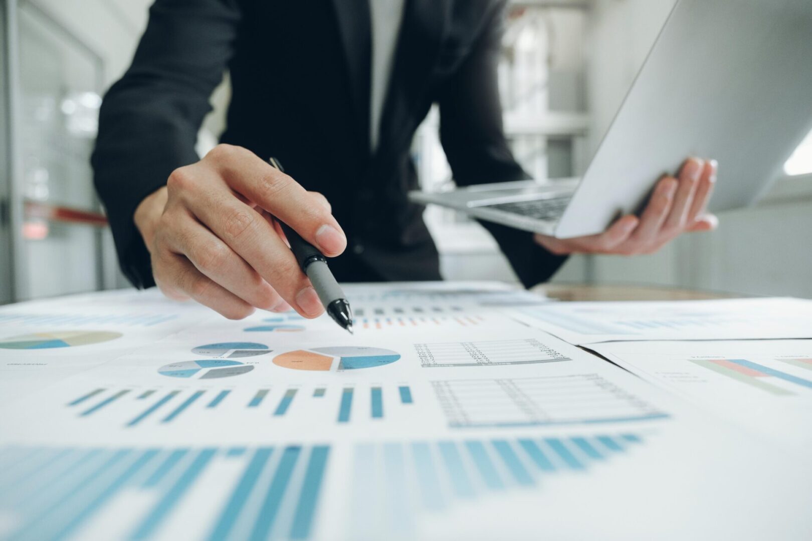 Person analyzing charts and graphs on paper while holding a laptop, using a pen to point at data. Business setting.