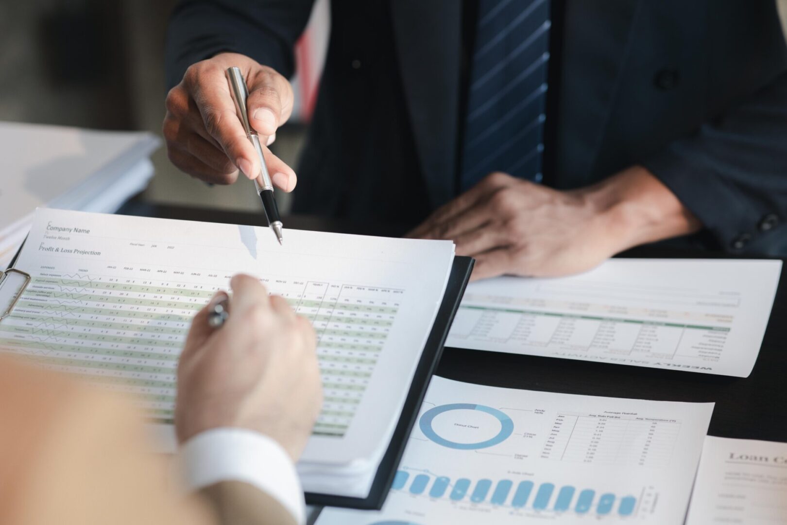 Two people in business attire reviewing financial documents with charts and graphs on a table. One person is pointing with a pen.