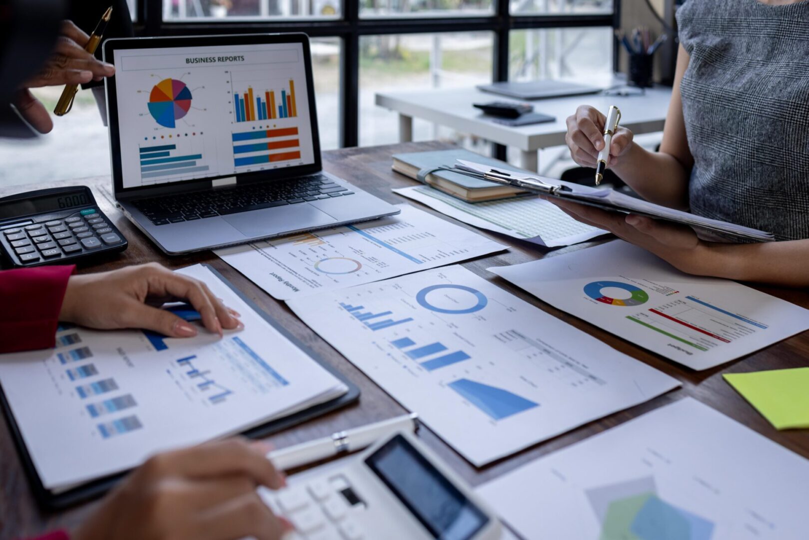 People working at a desk with business reports, graphs, and charts on paper and a laptop. One person writes on a clipboard.