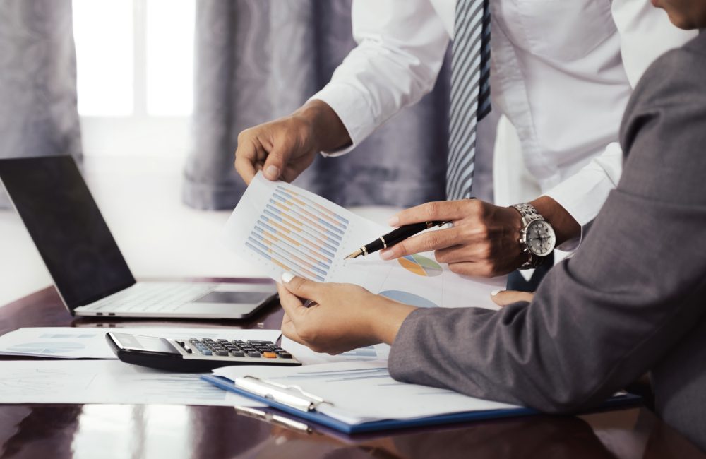 Two professionals in business attire review a document with graphs at a desk. A laptop, calculator, and clipboard are visible.